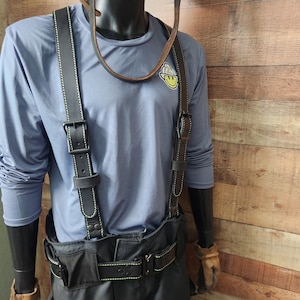 May include: A mannequin displays a work uniform, including a light blue long-sleeve shirt with a smiley face logo, black suspenders with green stitching, and a black belt. The outfit also features black pants and work gloves.
