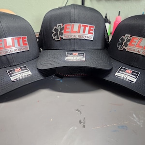 May include: Three black baseball caps with a structured front and a curved brim. Each cap features a silver and red logo that reads "ELITE MEDICAL RESPONSE." The caps have a Richardson 112 tag.