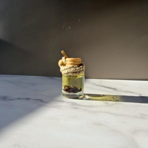 Könnte beinhalten: Eine kleine, durchsichtige Glasflasche, gefüllt mit einer hellgelben Flüssigkeit und getrockneten Pflanzenteilen am Boden. Die Flasche hat einen hellen Holzverschluss und ist mit einer dicken, cremefarbenen Kordel umwickelt.