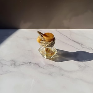 Könnte beinhalten: Eine kleine, klare, rautenförmige Glasflasche mit facettiertem Boden. Der Flaschenhals ist mit einer dicken, grauen, gesprenkelten Kordel umwickelt, gekrönt von einem runden, bernsteinfarbenen Holzstopfen und einer kleinen, passenden Perle.