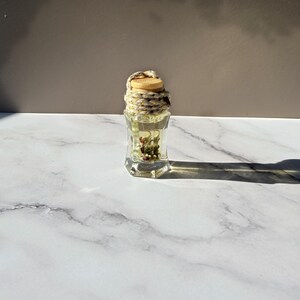 Könnte beinhalten: Eine kleine, durchsichtige, sechseckige Glasflasche, gefüllt mit einer hellen Flüssigkeit und getrockneten botanischen Elementen. Die Flasche ist mit einem hellen Holzstopfen verschlossen, der mit einem dicken, cremefarbenen Seil mit Goldsprenkeln umwickelt ist.