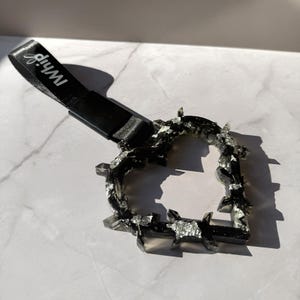 May include: A black heart-shaped handle with a barbed wire design, featuring embedded silver foil accents. The handle is attached to a black fabric strap with the white text "Whip" printed on it, designed for use as a vehicle grab handle.