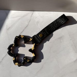 May include: A black flower-shaped ring with gold leaf accents attached to a black textured strap featuring a crocodile-skin pattern and a single brass-colored metal snap button. The object is designed with a hollow center and a smooth finish.