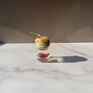 Könnte beinhalten: Eine kleine, durchsichtige Glasflasche gefüllt mit einer klaren Flüssigkeit und einem einzelnen rosa Blütenblatt. Die Flasche hat einen hellen Holzverschluss und ist mit einer dicken, gesprenkelten beigen Kordel umwickelt, die als Schlaufe dient.
