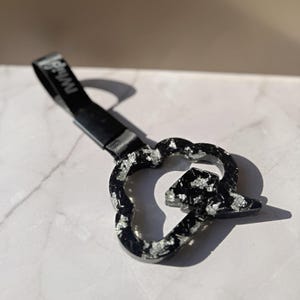 May include: A black resin handle with silver foil flakes in a cloud shape, featuring a lightning bolt cutout. A black strap is attached to the top, with the text "iWhip" printed in white on the strap. The object has a glossy, reflective finish.