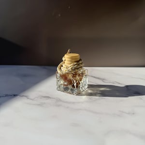 Könnte beinhalten: Eine kleine, quadratische Glasflasche mit einer facettierten, strukturierten Außenfläche. Die Flasche ist mit einem hellen Holzverschluss versehen und hat eine dicke, geflochtene Kordel um den Hals, die zum Aufhängen oder Präsentieren gedacht ist.