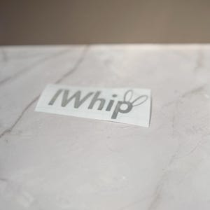 Puede incluir: Una calcomanía rectangular con el texto "IWhip" en una fuente negrita de color gris oscuro sin remates. La letra "p" al final de la palabra está estilizada con dos bucles alargados de forma ovalada que se extienden hacia arriba, como orejas de conejo.