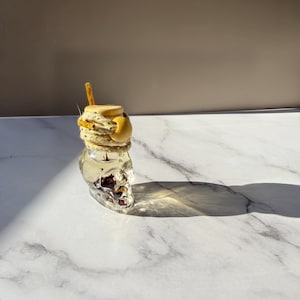 Könnte beinhalten: Eine durchsichtige, schädelförmige Glasflasche, gefüllt mit einer Flüssigkeit, die dunkle botanische Elemente enthält. Der Flaschenhals ist mit einer dicken, strukturierten beigen Kordel mit Goldsprenkeln umwickelt, die durch eine gelbe Holzperle fixiert ist.