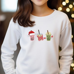 Może przedstawiać: Biała bluza z nadrukiem trzech kubków z bubble tea w zimowych dodatkach. Kubki z bubble tea noszą czapkę Świętego Mikołaja, szalik i rękawiczki.