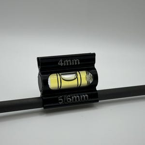 May include: A black level tool with a clear, yellow-tinted vial containing a bubble, mounted on a dark rod. The tool is marked with "4mm" and "5/6mm" in white text. The background is plain white.