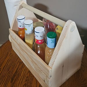 May include: A wooden caddy with a handle, filled with various bottles of sauces and a roll of paper towels. The caddy is made of unfinished wood and is designed for easy transport of condiments and supplies.