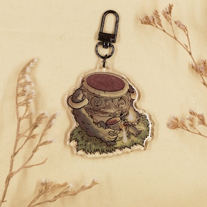 Könnte beinhalten: Ein klarer Acryl-Schlüsselanhänger mit einer Cartoon-Illustration eines pelzigen Wesens, das einen dekorativen Topf hält. Der Topf ist braun mit komplizierten Mustern. Der Schlüsselanhänger hat einen schwarzen Verschluss und befindet sich auf einem beigen Hintergrund mit getrockneten Blumen.