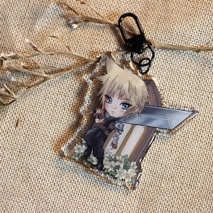 Könnte beinhalten: Ein durchsichtiger Acryl-Schlüsselanhänger mit einer Chibi-Illustration eines blonden Charakters, der ein Schwert hält. Der Charakter steht auf einem Bett aus weißen Blumen. Der Schlüsselanhänger hat einen schwarzen Metallring.