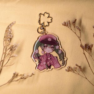 May include: A clear acrylic charm featuring a cartoon character in a pink outfit and hat, adorned with floral accents. The charm has a gold-colored keychain attachment. The character has green hair and is smiling. The background is a light yellow color.