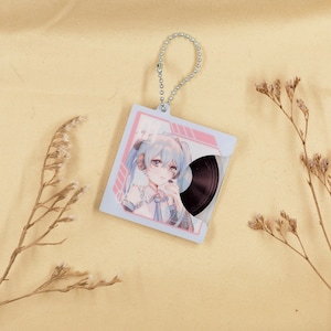 May include: A square keychain featuring an anime-style illustration of a girl with blue hair and a vinyl record design. The keychain has a silver chain and a light blue and pink border.