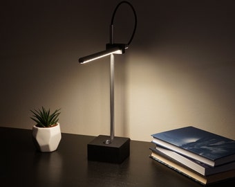 May include: A silver desk lamp with a black base and a flexible arm. The lamp is on a black surface with a small potted plant and a stack of books.