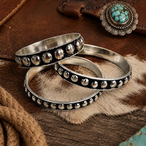 Op de afbeelding: Drie zilverkleurige armbanden met een zwarte inlegband versierd met een herhalend patroon van kleine, gepolijste zilverkleurige metalen bolletjes. De armbanden hebben een glad, reflecterend binnenoppervlak en een consistente ronde vorm.