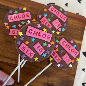 Puede incluir: Cuatro piruletas de chocolate con el nombre "CHLOE" y las palabras "SIX IS" en letras rosas. El chocolate está decorado con chispas de estrellas de colores. Las piruletas están sobre palitos blancos y descansan sobre una superficie de madera.