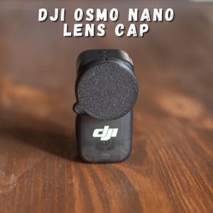 Puede incluir: Se muestra una tapa de lente negra DJI Osmo Nano, que protege la lente de la cámara. El texto "DJI OSMO NANO LENS CAP" se muestra en la parte superior. La cámara es negra con el logotipo de DJI.
