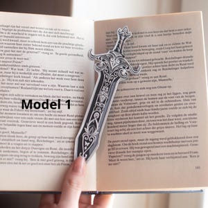 May include: A black and silver sword-shaped bookmark with ornate detailing. The bookmark is placed on an open book, with the tip of the sword pointing towards the text.