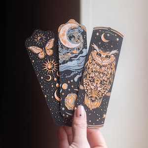 Op de afbeelding: Drie zwarte bladwijzers met hemelse ontwerpen. Eén heeft een vlinder, zon en halve manen. Een andere heeft een maan en wolken. De derde bladwijzer toont een uil met een halve maan en sterren. De ontwerpen zijn in goud en lichtblauw.