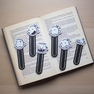 May include: Six character bookmarks in black and white are displayed on an open book. Each bookmark has a different cartoon face. The bookmarks are made of a black material with a white face design.