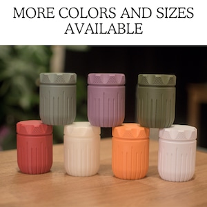 Puede incluir: Siete pequeños contenedores con lados acanalados en varios colores, incluyendo verde, morado, rojo, blanco y naranja. Los contenedores están dispuestos en forma de pirámide sobre una superficie de madera. El texto "MÁS COLORES Y TALLAS DISPONIBLES" se muestra sobre los contenedores.