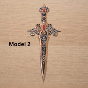 May include: Ornate dagger with a black blade and gold and silver filigree detailing. The hilt features red gemstone accents. The text "Model 2" is printed in black on the left side of the image. The dagger is displayed on a light brown wooden surface.