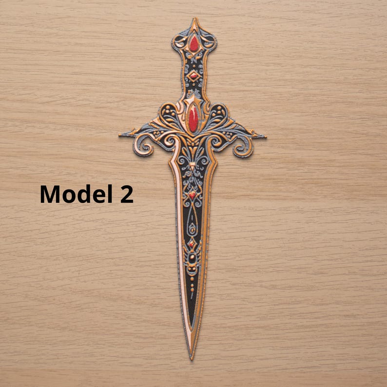 May include: Ornate dagger with a black blade and gold and silver filigree detailing. The hilt features red gemstone accents. The text "Model 2" is printed in black on the left side of the image. The dagger is displayed on a light brown wooden surface.