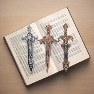 May include: Three decorative sword-shaped bookmarks rest on an open book. The bookmarks feature intricate designs with silver, gold, and black finishes. One has a red gem. The book's pages are visible, creating a literary theme.