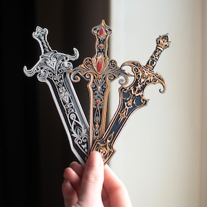 May include: Three ornate sword-shaped bookmarks, one silver, one black with gold accents and red jewels, and one black with gold accents.