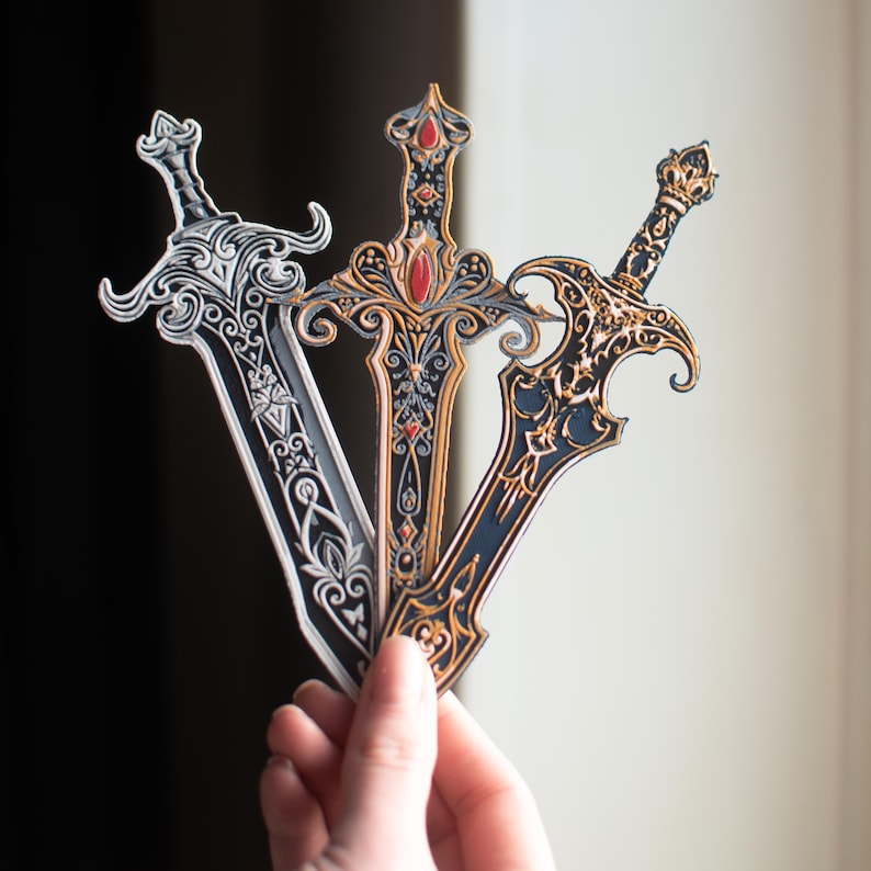 May include: Three decorative swords with ornate designs. One sword is silver and black, another is gold and black with a red gem, and the third is gold and navy blue. The swords have detailed handles and blades.