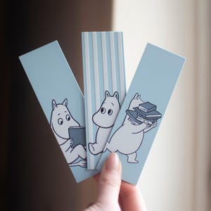 Op de afbeelding: Drie lichtblauwe bladwijzers met illustraties van Moomin-personages. Eén bladwijzer heeft witte verticale strepen. De Moomin-personages zijn in zwart en wit, met één die een boek leest en een ander die een stapel boeken draagt.