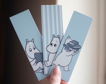 May include: Three light blue bookmarks featuring illustrations of Moomin characters. One bookmark has vertical white stripes. The Moomin characters are depicted in black and white, with one reading a book and another carrying a stack of books.
