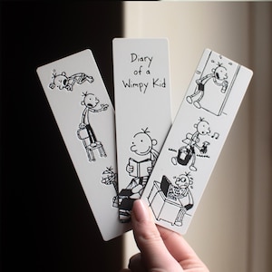 Puede incluir: Tres marcapáginas blancos con ilustraciones en blanco y negro de "Diario de Greg". Los marcapáginas muestran escenas de dibujos animados de un niño. El texto "Diary of a Wimpy Kid" está impreso en un marcapáginas.