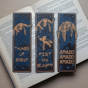 Puede incluir: Tres marcapáginas rectangulares con un fondo azul oscuro y un diseño temático espacial. Cada marcapáginas presenta una frase diferente: "THUMBS UP BABY?", "FIST MY BUMPY?" y "AMAZE! AMAZE! AMAZE!".