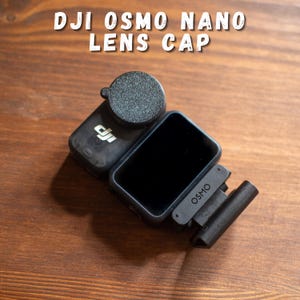 Può includere: Fotocamera DJI Osmo Nano nera con un copriobiettivo, appoggiata su una superficie di legno. Il copriobiettivo è rotondo e strutturato. Il testo "DJI OSMO NANO LENS CAP" è visualizzato nella parte superiore dell'immagine.