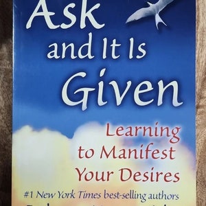 Puede incluir: Una portada de libro con un fondo de cielo azul y un pájaro blanco volando. El título es "Ask and It Is Given: Learning to Manifest Your Desires." El subtítulo es "#1 New York Times best-selling authors Esther and Jerry Hicks The Teachings of Abraham."