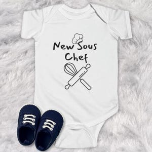 May include: White baby onesie with black text that reads "New Sous Chef" and a chef's hat, whisk, and rolling pin graphic.
