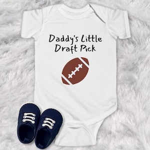 May include: White baby onesie with the text "Daddy's Little Draft Pick" and a brown football graphic. Includes two navy blue baby shoes with white laces. Perfect for a baby shower or a new baby gift.