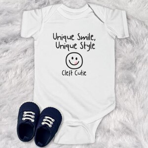 May include: White baby onesie with black text that reads "Unique Smile, Unique Style" and a smiley face with a pink tongue. Below the smiley face is the text "Cleft Cutie".