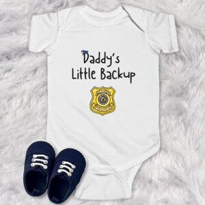 May include: White baby onesie with the text "Daddy's Little Backup" and a cartoon police badge.
