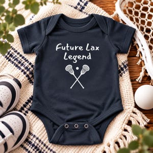May include: A dark navy blue short-sleeved baby bodysuit featuring the white printed text Future Lax Legend above a graphic of two crossed lacrosse sticks and a small ball. The garment has a round neckline and three metal snaps at the bottom.