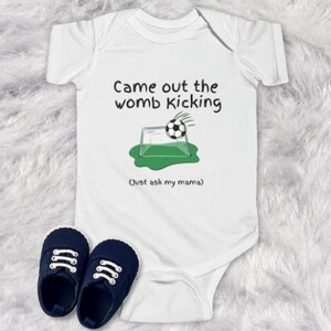 May include: White baby onesie with black text that reads "Came out the womb kicking. Just ask my mama.". The onesie features a cartoon illustration of a soccer ball kicking a goal.