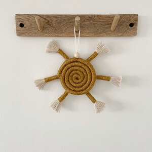 May include: A mustard yellow sun-shaped wall hanging with a spiral design and white fringe. The sun is hanging from a wooden hook.