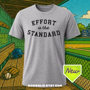 May include: A heather grey short-sleeve t-shirt featuring the black text "EFFORT is the STANDARD" printed on the chest. The shirt has a crew neckline and a standard fit. A small green tag with the word "New" is visible in the bottom right corner.