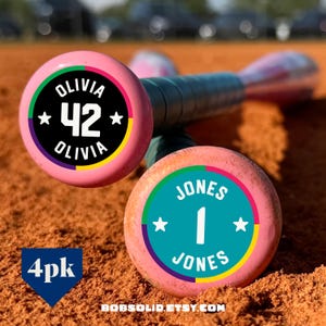 May include: Two pink baseball bats with circular end caps. One cap features a black background with the white text OLIVIA and the number 42. The other cap has a teal background with the white text JONES and the number 1. Both have colorful borders.
