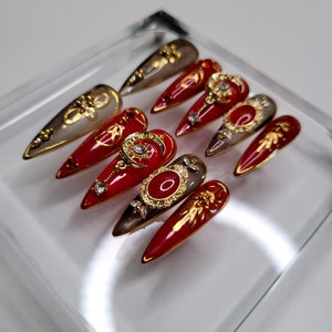 May include: Set of ten red and gold false nails with intricate designs. Each nail features a different design, including a crescent moon, a sun, and a palm tree. The nails are arranged in a glass display case.