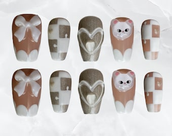 May include: A set of ten artificial nails in various designs. The nails feature a mix of colors including brown, white, and silver. Some nails have bow accents, puzzle piece patterns, heart shapes, and a cat face design. The nails are oval-shaped.