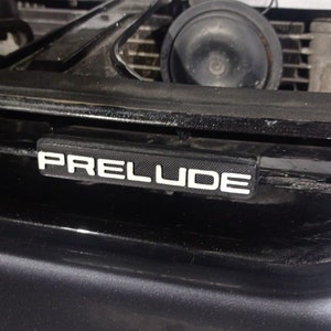 Peut inclure: Gros plan sur une calandre de voiture noire avec un emblème "PRELUDE" en plastique noir avec des lettres blanches.